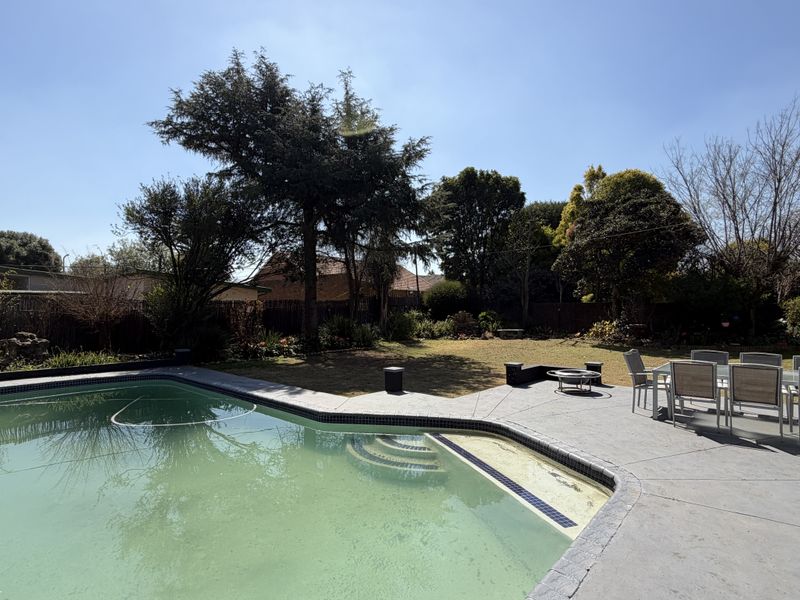 Outdoor entertainment area featuring a swimming pool, concrete patio, and a spacious garden with mature trees and perimeter fencing.