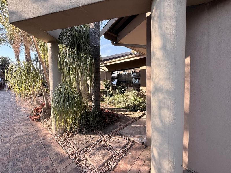 Front entrance area featuring concrete columns, paved driveway, and established garden beds with casement windows visible on the building facade.