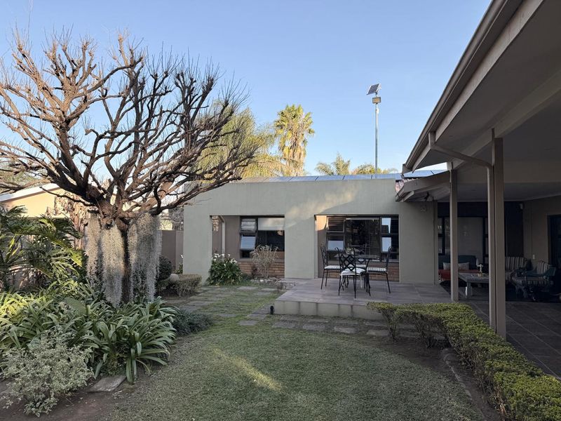 A well-maintained rear garden featuring a paved patio area, established trees, and a covered walkway along the side of the house.