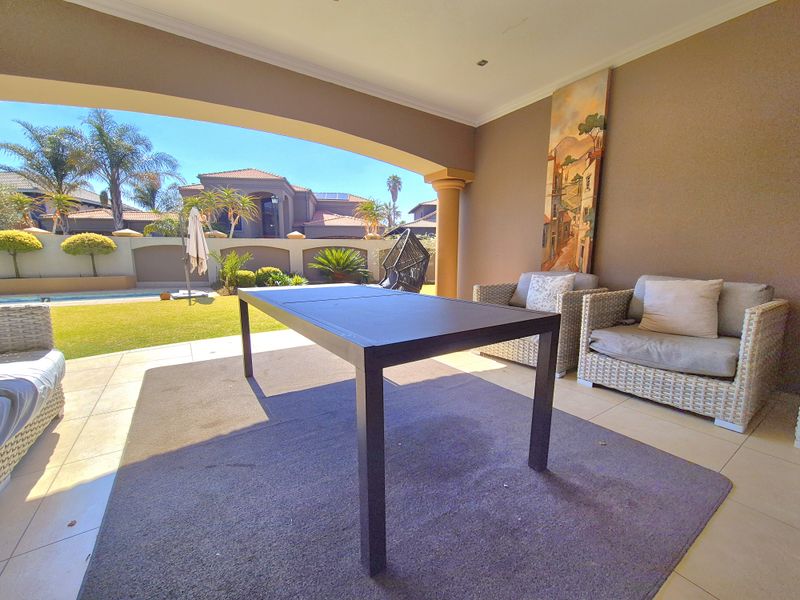 A spacious covered patio featuring tiled flooring and recessed lighting, overlooking a landscaped garden and swimming pool area.
