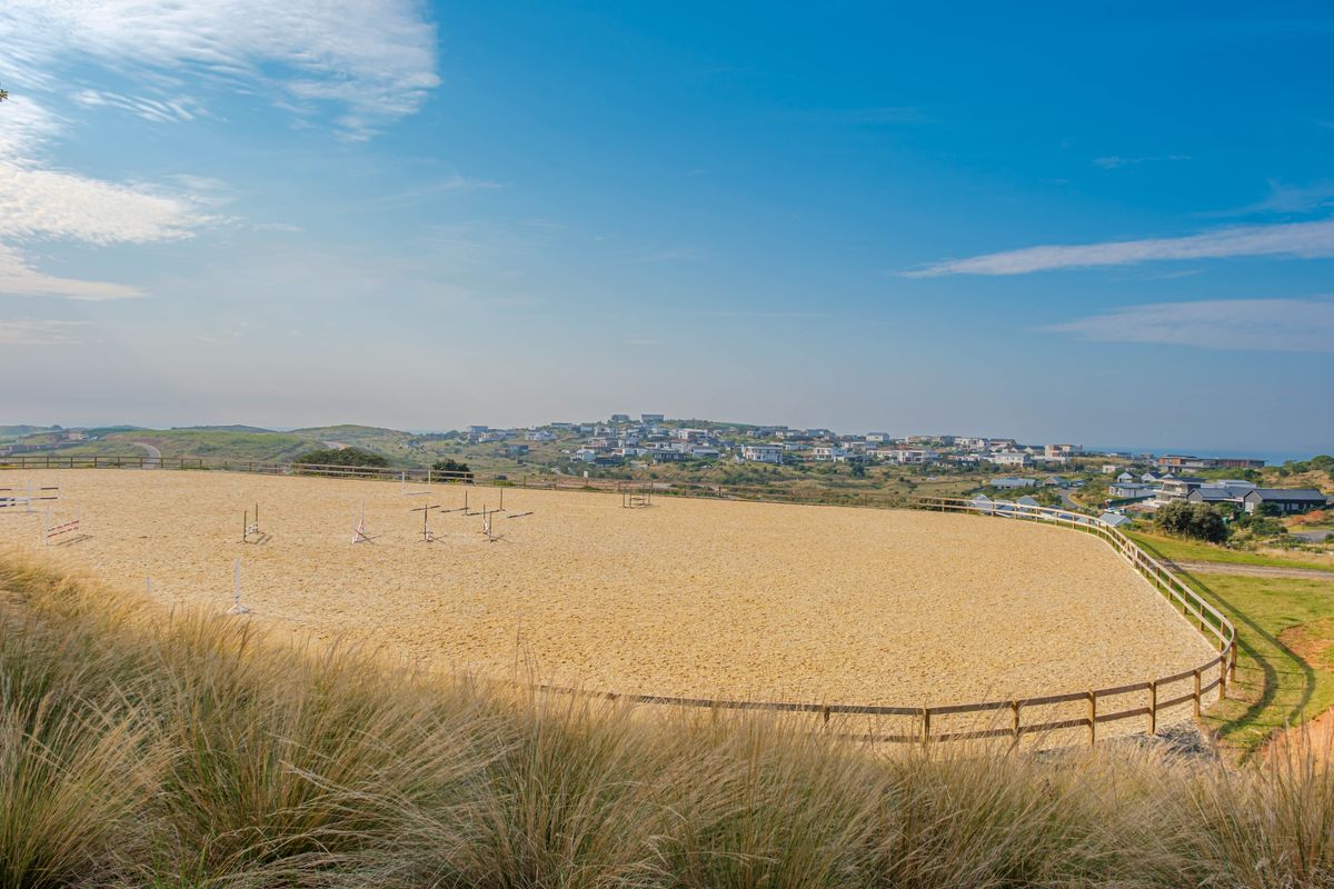 A well-maintained outdoor riding arena with a sand surface and timber post and rail fencing, set against a scenic coastal landscape.