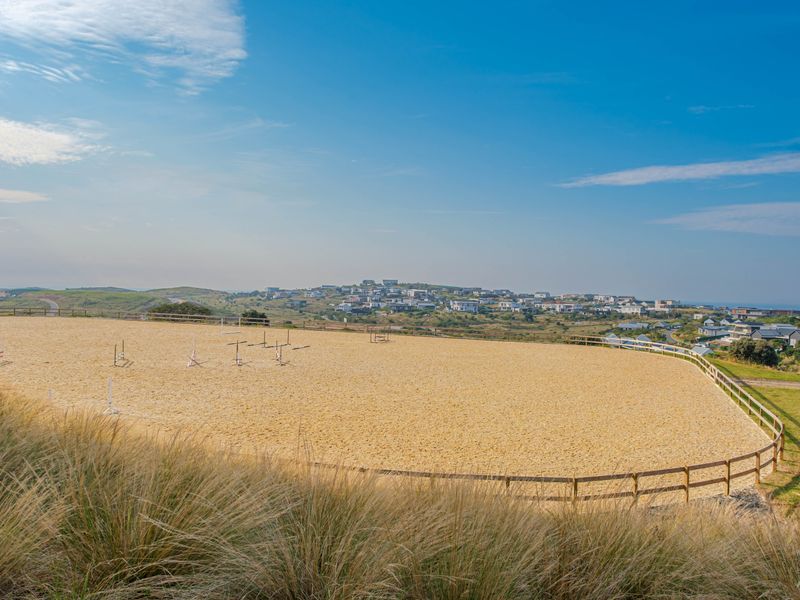 A well-maintained outdoor riding arena with a sand surface and timber post and rail fencing, set against a scenic coastal landscape.