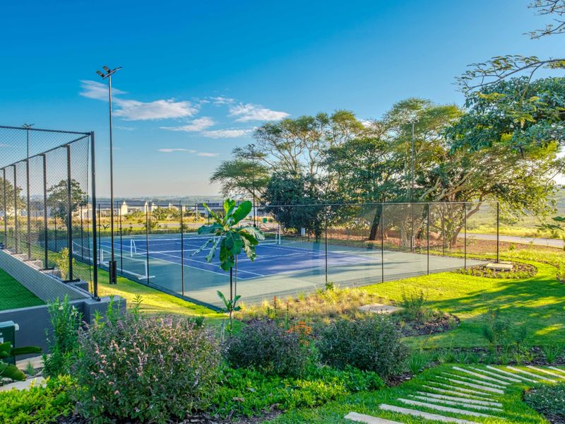 A well-maintained outdoor tennis court featuring perimeter fencing, floodlighting, and surrounding landscaped gardens with stone pathways.