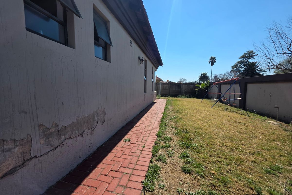 Rear exterior view of the property featuring a brick paved walkway, lawn area, and boundary wall, showing visible signs of exterior wall plaster damage.