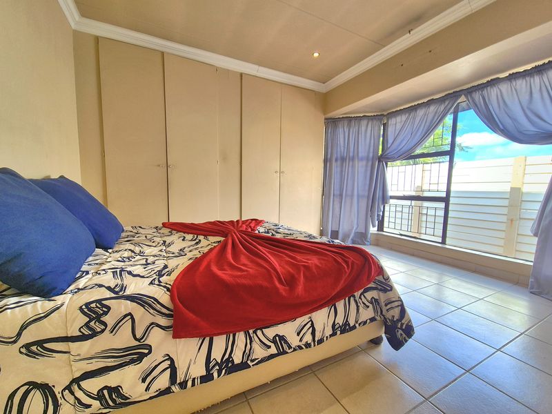 A bedroom featuring built-in cupboards, tiled flooring, and large windows with security bars, offering a functional space with potential for cosmetic updates.