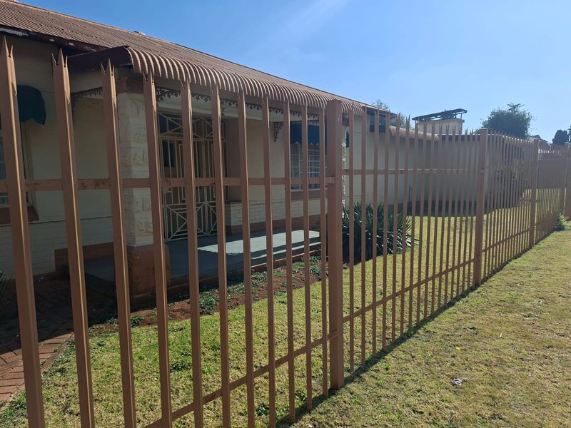 Front view of a house featuring a corrugated metal roof, brick facade, and secure metal perimeter fencing.