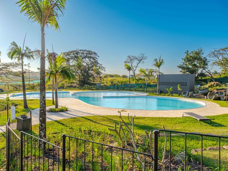 A well-maintained outdoor swimming pool area featuring a paved deck, landscaped garden surroundings, and secure metal perimeter fencing.