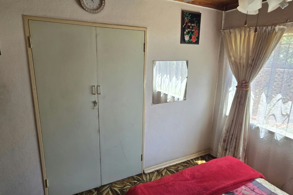 A bedroom featuring a built-in wardrobe, ceiling fan, and security bars on the window. The room requires cosmetic updates to modernize the finishes.