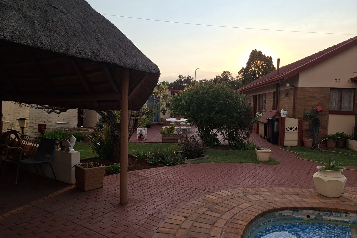 A rear garden area featuring a thatched lapa, brick paved patio, and a swimming pool, set against the brick exterior of the house.
