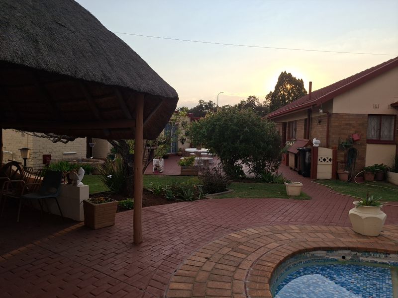 A rear garden area featuring a thatched lapa, brick paved patio, and a swimming pool, set against the brick exterior of the house.
