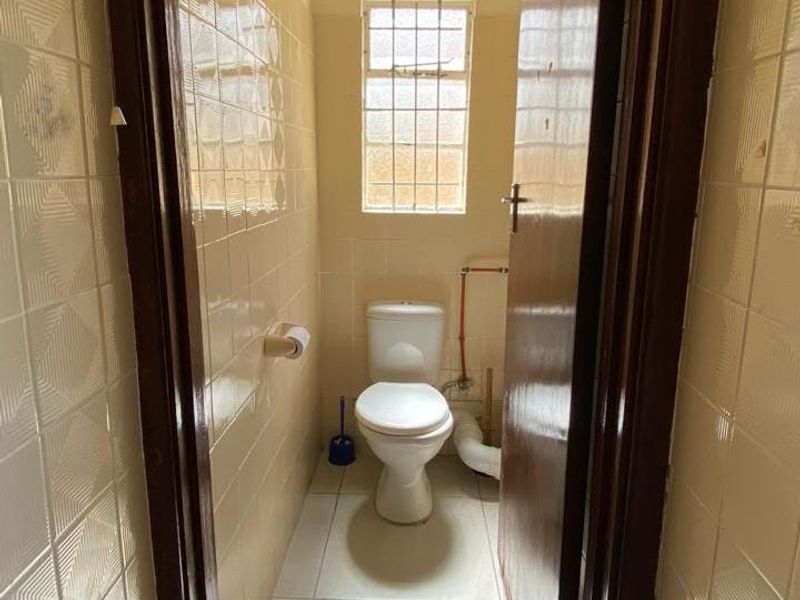 A compact, dated bathroom featuring ceramic wall and floor tiles, a standard toilet, and exposed plumbing, requiring modernization.