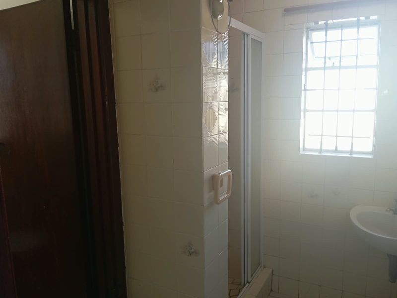 A dated bathroom featuring a shower enclosure, wall-mounted basin, and tiled walls with security bars on the window.
