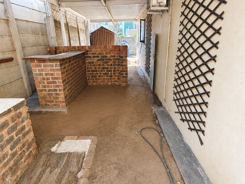 Covered outdoor patio area featuring a built-in brick bar, corrugated roof, and trellis wall, currently requiring flooring installation and general maintenance.