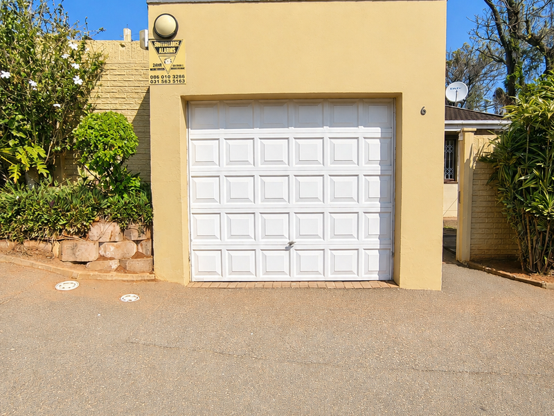 A single-car garage with a white sectional door, exterior lighting, and security alarm signage, situated at the front of the townhouse property.