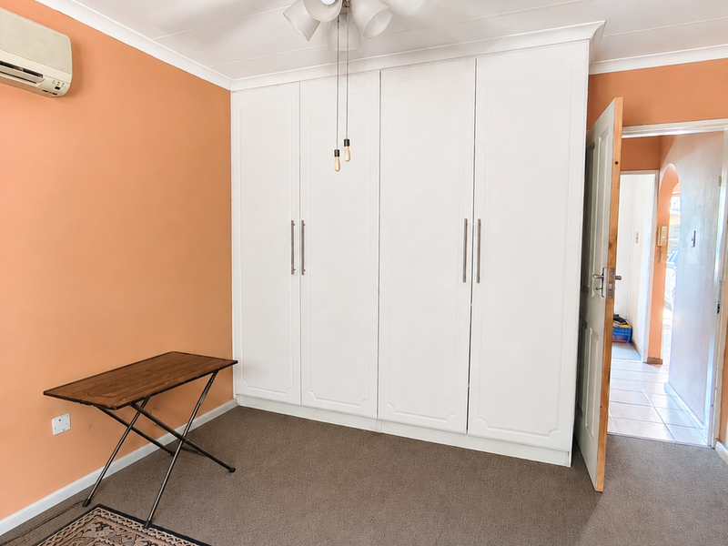 A bedroom featuring built-in wardrobes, a ceiling fan, and a wall-mounted air conditioning unit, with neutral carpet flooring and orange wall paint.