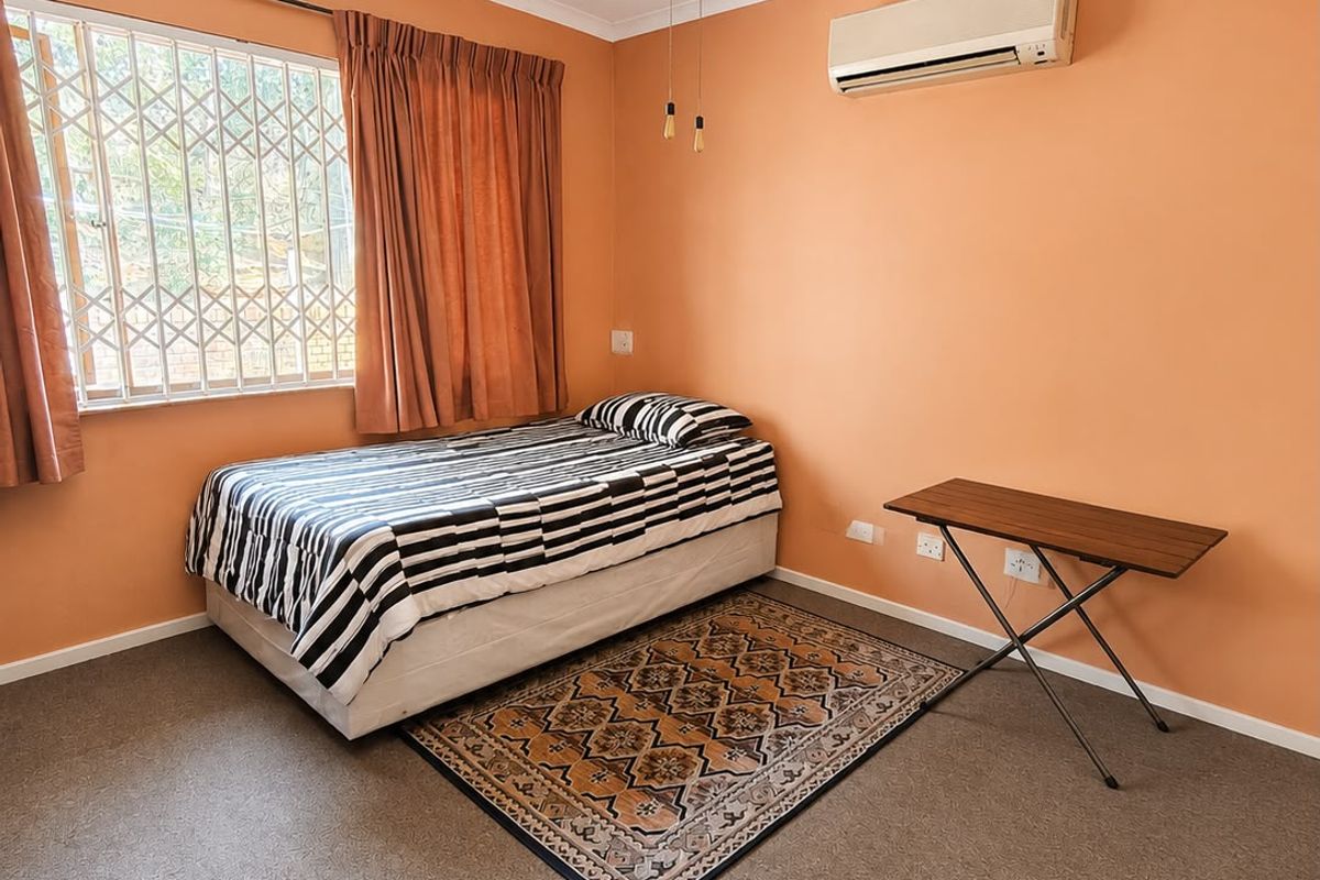 A functional bedroom featuring carpeted flooring, a ceiling fan, and a wall-mounted air conditioning unit, with security bars installed on the window.