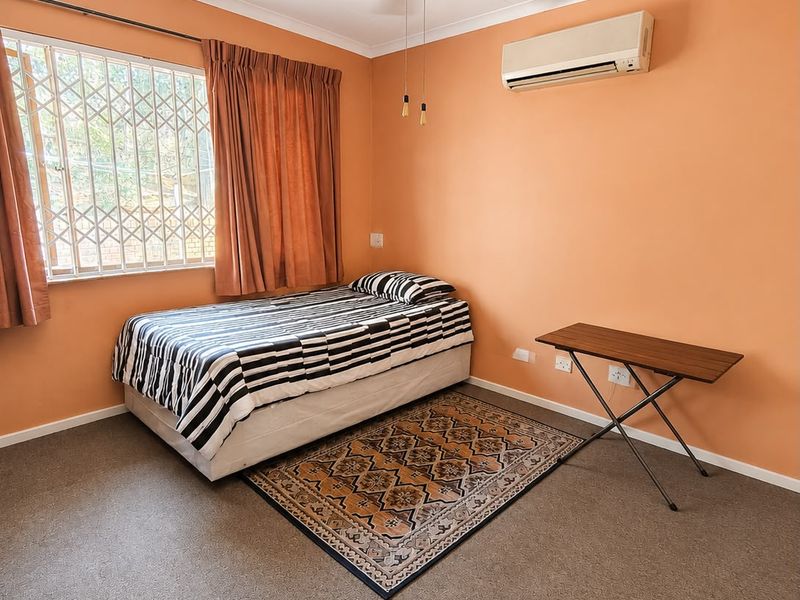 A functional bedroom featuring carpeted flooring, a ceiling fan, and a wall-mounted air conditioning unit, with security bars installed on the window.