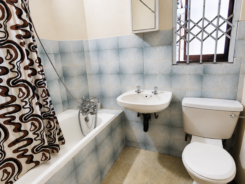 A functional bathroom featuring a bathtub, wall-mounted basin, and toilet, finished with dated wall tiling and a security-gated window.