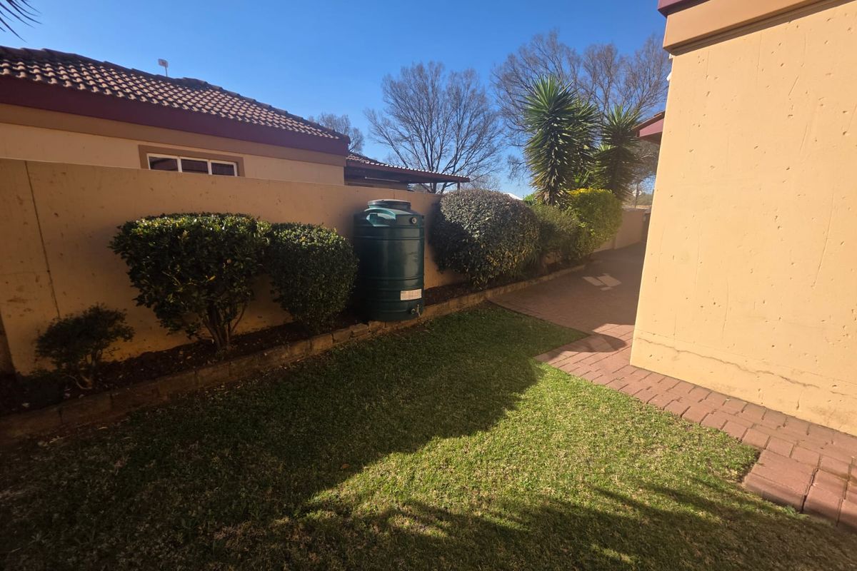 A rear exterior view of the townhouse featuring a small lawn, paved pathway, boundary wall, and a green water storage tank.