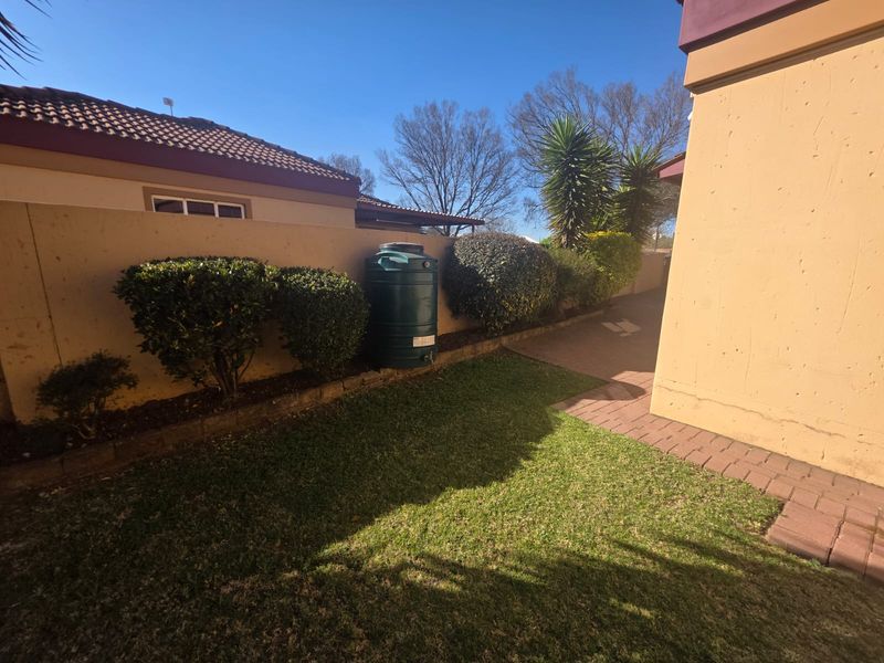 A rear exterior view of the townhouse featuring a small lawn, paved pathway, boundary wall, and a green water storage tank.