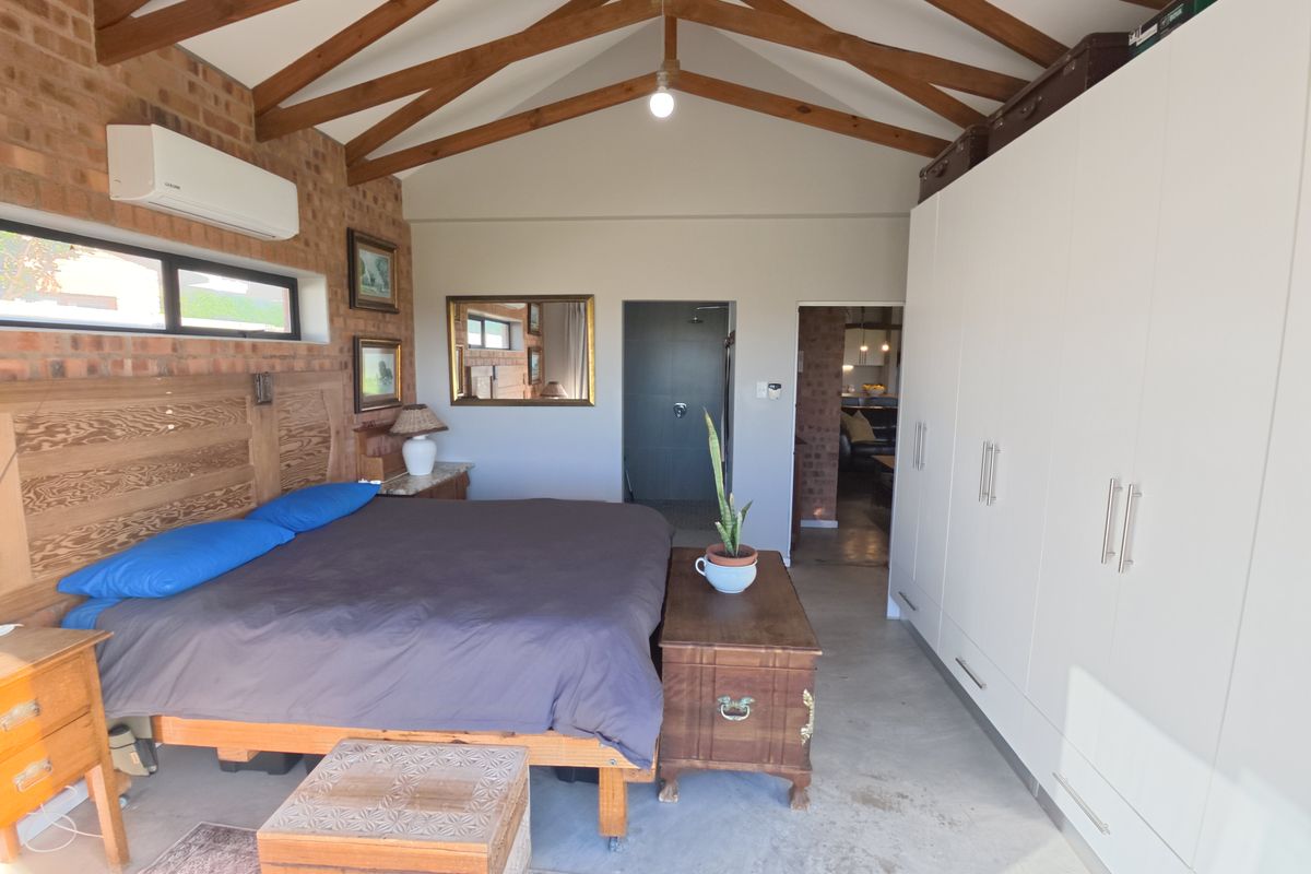 Spacious bedroom featuring exposed timber roof trusses, polished concrete floors, exposed brick feature wall, built-in cupboards, and an en-suite bathroom.