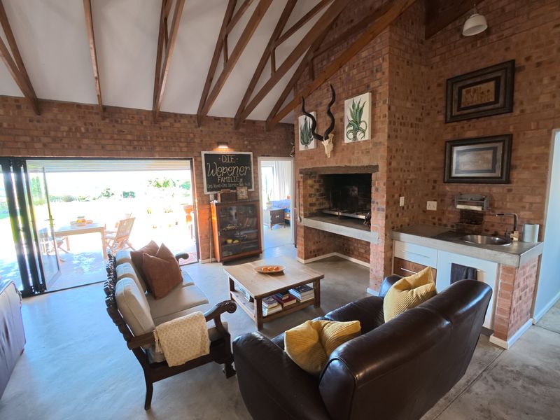 Spacious open-plan living area featuring exposed timber rafters, a large brick fireplace with built-in braai, and seamless access to the outdoor patio through bi-fold doors.