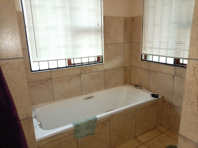 A bathroom featuring a built-in bathtub with tiled surround, tiled walls, and windows fitted with venetian blinds and security bars.