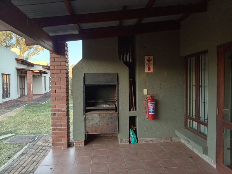 Covered patio area featuring a built-in braai, tiled flooring, and exposed timber rafters, overlooking the garden space.