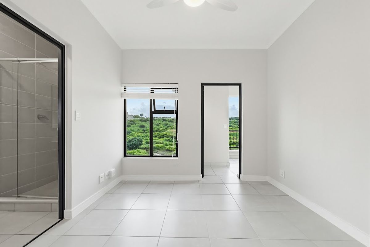 A bright, modern, and empty bedroom featuring neutral tiled flooring, a ceiling fan, and an en-suite bathroom with a view of the surrounding landscape.