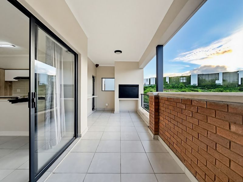 A spacious covered balcony featuring tiled flooring, a built-in braai, and a brick balustrade, offering a clean and modern outdoor entertainment space.