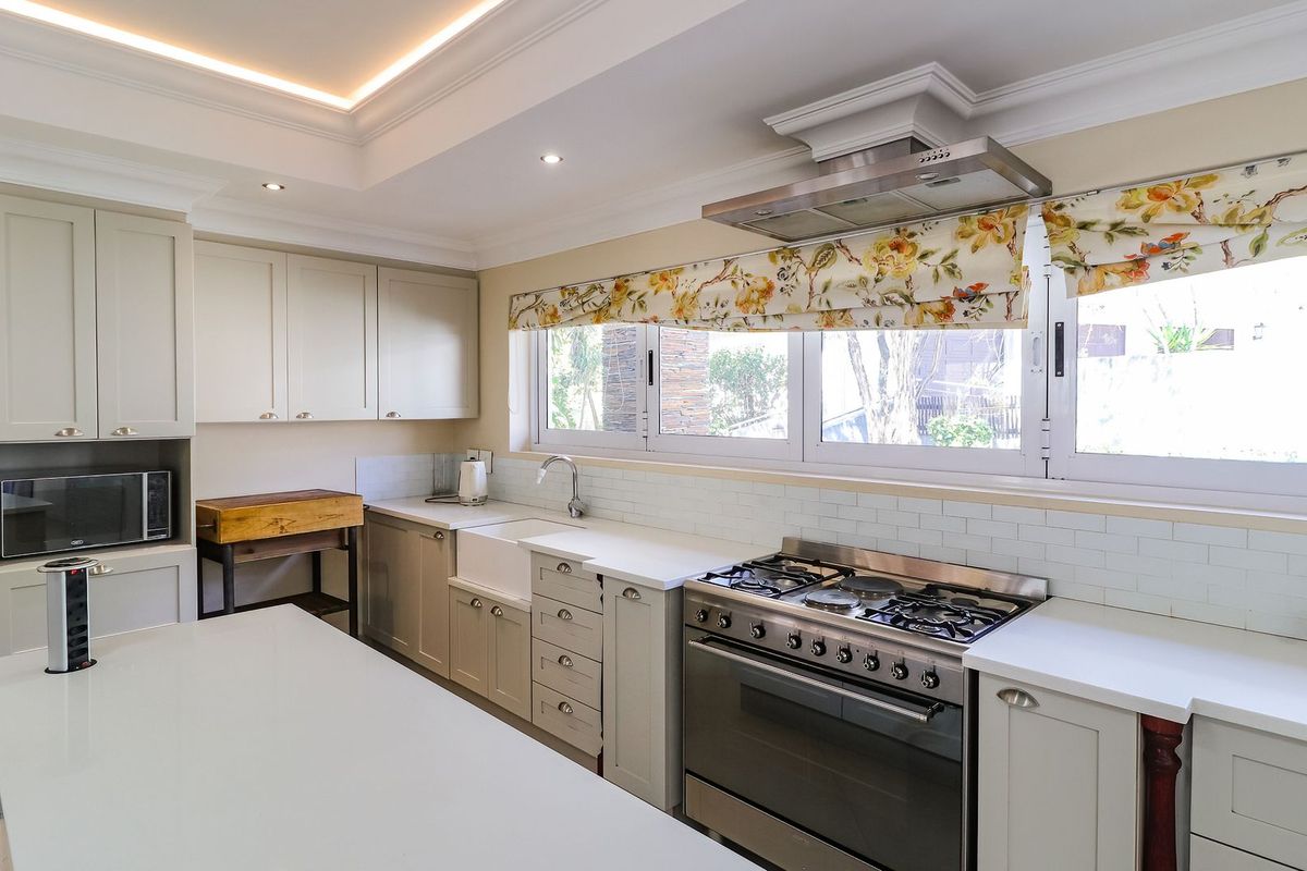Modern kitchen featuring light-toned cabinetry, stone countertops, a gas stove with extractor, apron sink, and elegant cove lighting.