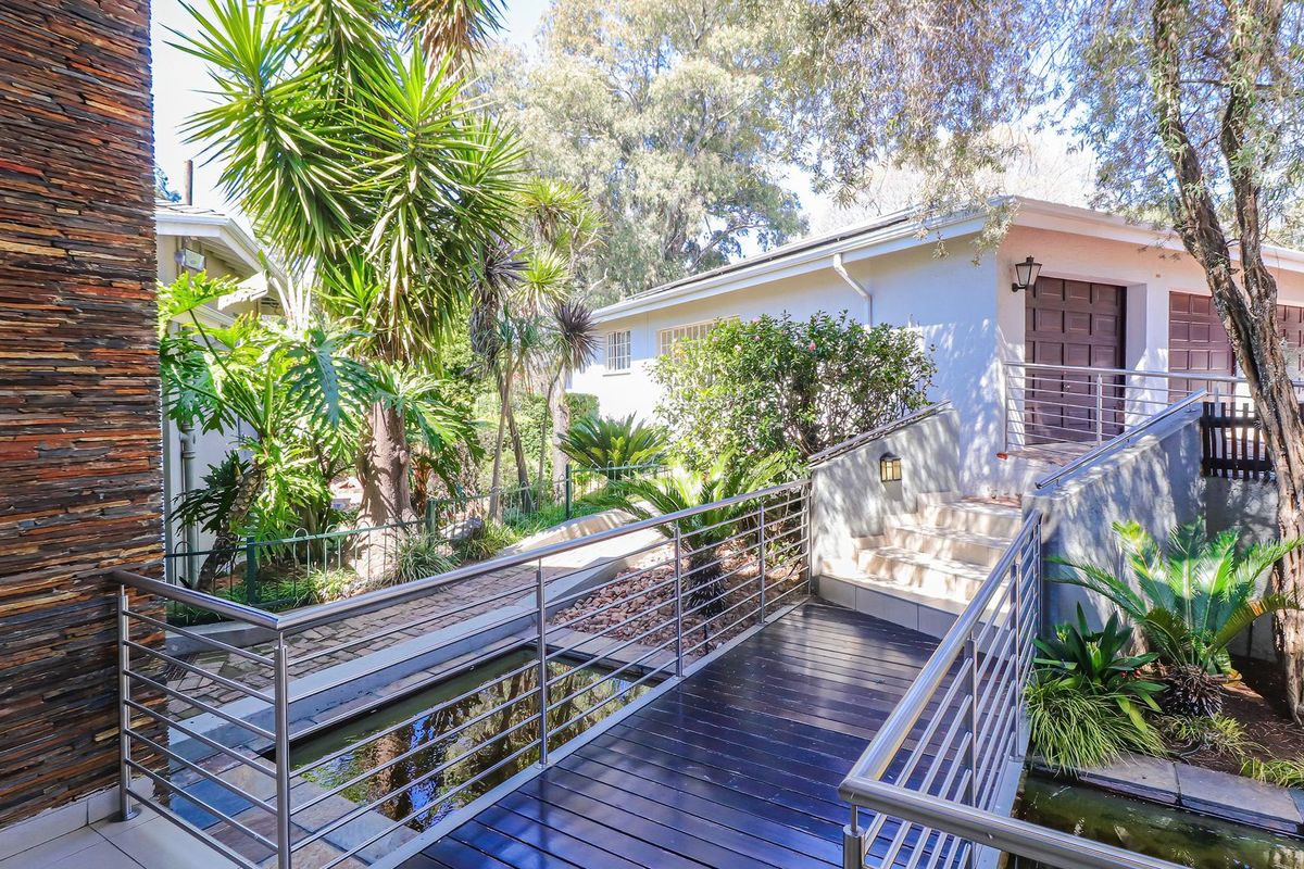 A landscaped rear garden area featuring a timber bridge over a water feature, stone-clad architectural elements, and modern stainless steel balustrades.