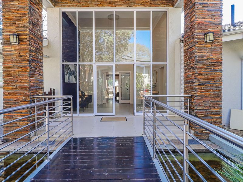 Grand entrance featuring a timber footbridge over a water feature, flanked by stone-clad pillars and a striking floor-to-ceiling glass facade.