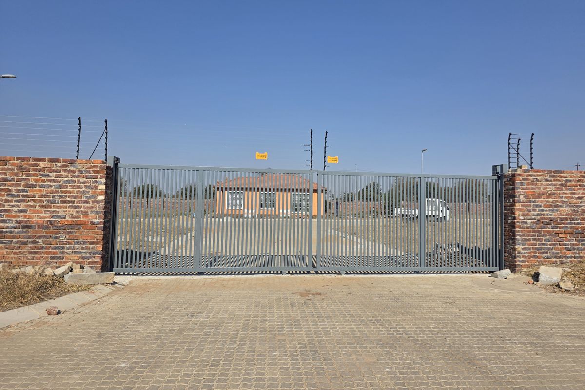 Front view of the property featuring a secure metal sliding gate, brick perimeter wall with electric fencing, and a paved driveway entrance.
