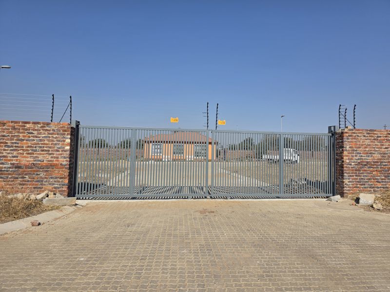 Front view of the property featuring a secure metal sliding gate, brick perimeter wall with electric fencing, and a paved driveway entrance.