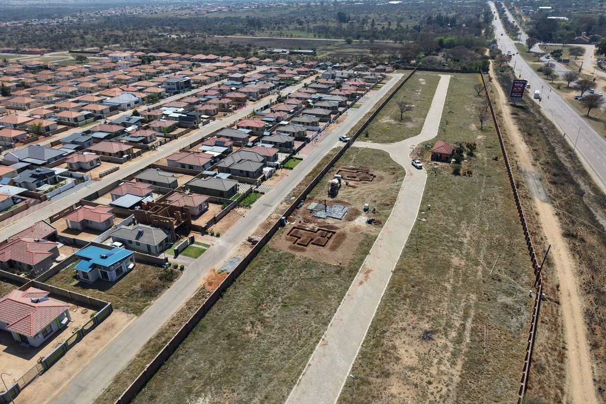 Aerial view of a residential development site showing paved access roads, perimeter fencing, and active foundation excavations for new housing units.