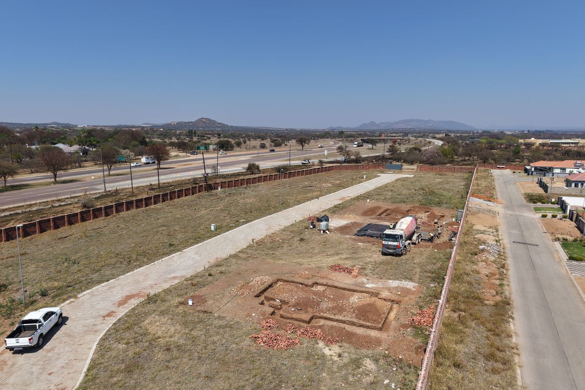 Aerial view of a vacant residential plot featuring an excavated foundation, paved driveway, and perimeter brick walling, situated near a main road.