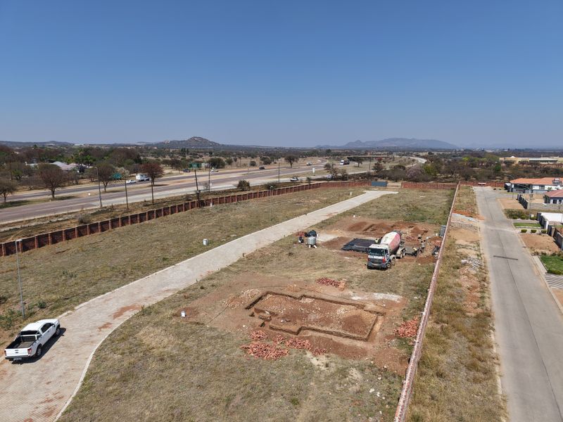 Aerial view of a vacant residential plot featuring an excavated foundation, paved driveway, and perimeter brick walling, situated near a main road.