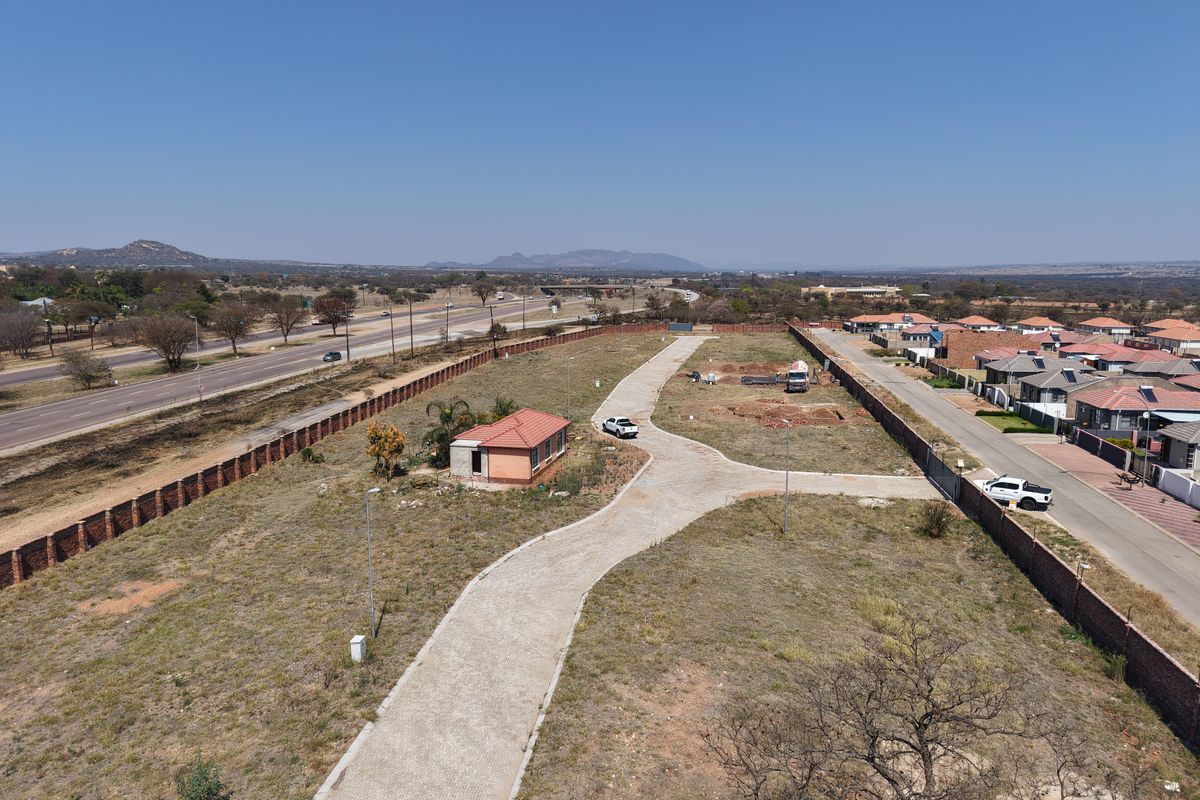 Aerial view of a vacant residential plot featuring a paved driveway, perimeter walling, and a small existing structure, situated near a main road.