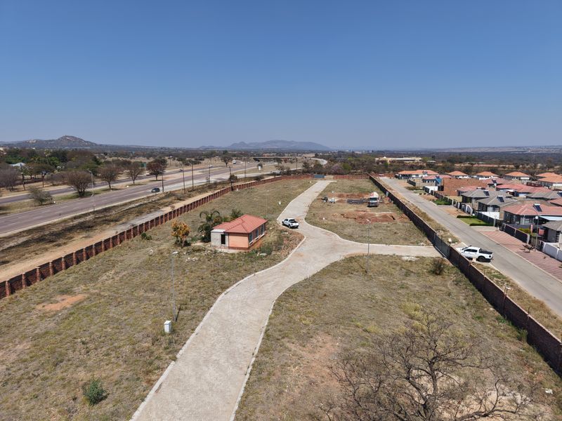 Aerial view of a vacant residential plot featuring a paved driveway, perimeter walling, and a small existing structure, situated near a main road.