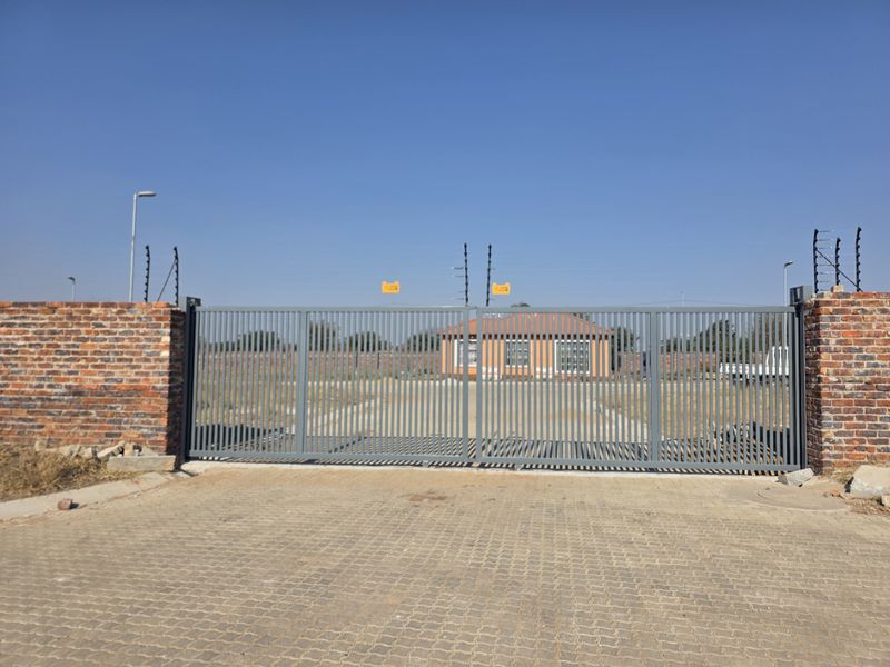 Front view of a residential property featuring a secure sliding metal gate, brick perimeter wall with electric fencing, and a paved driveway entrance.