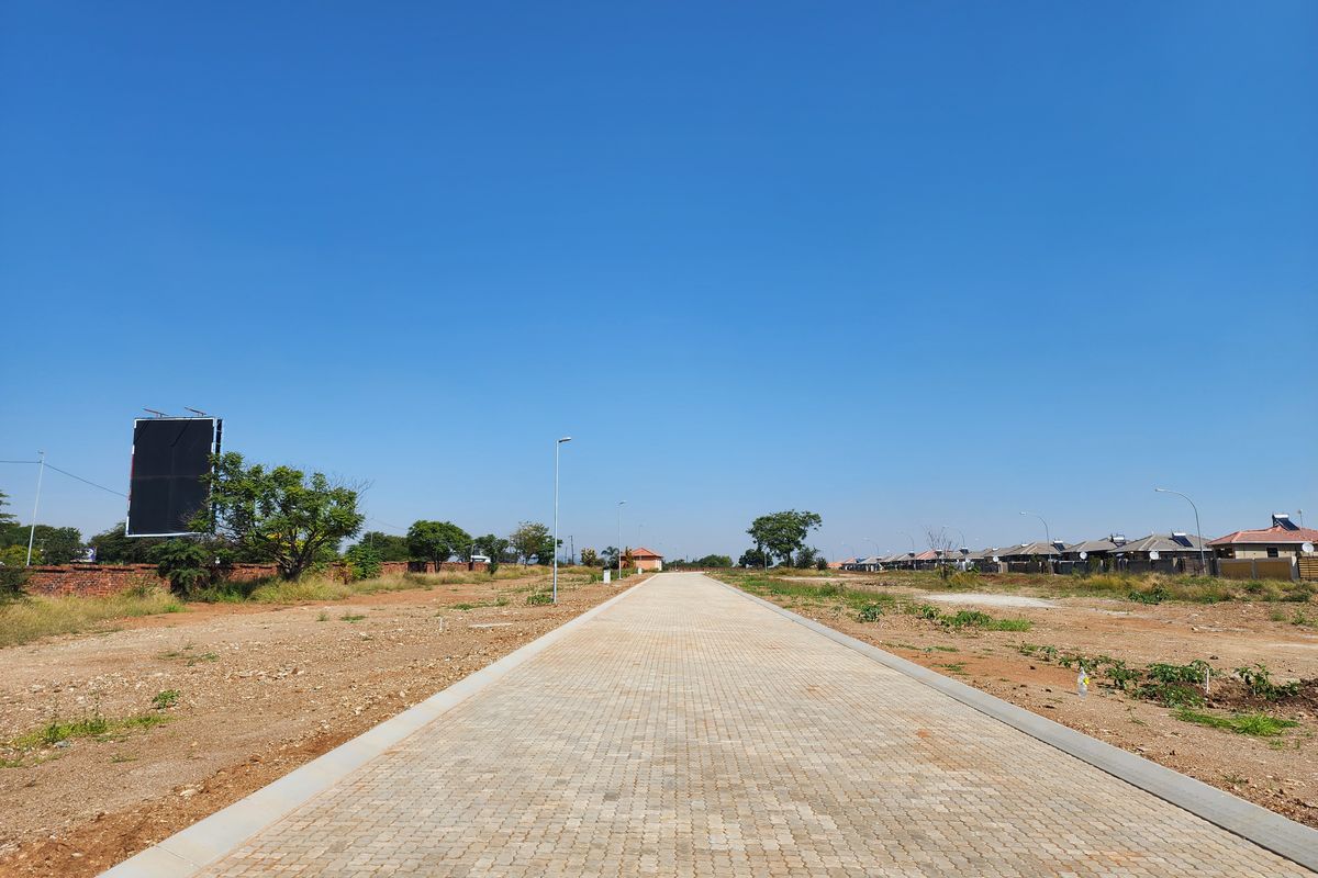 A clear view of a paved residential street within a developing estate, featuring established street lighting and surrounding vacant land plots.