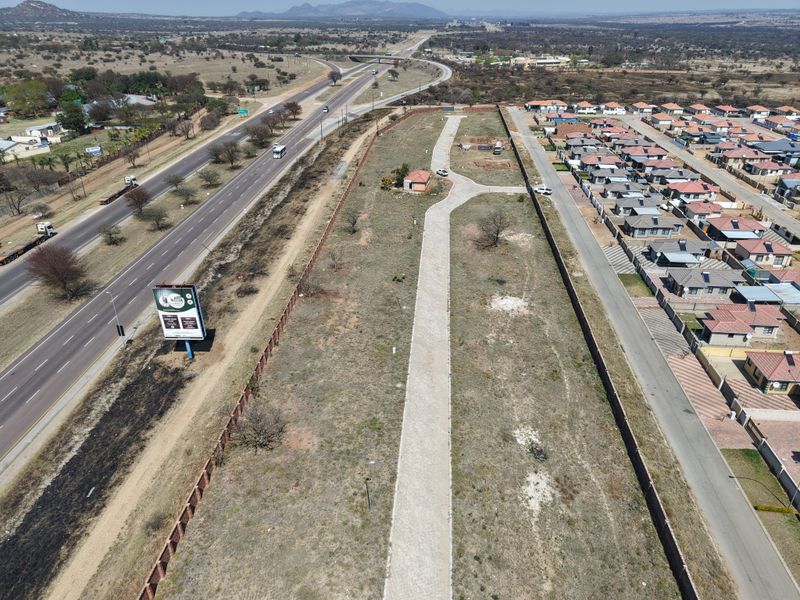 Aerial view of a vacant residential plot featuring a paved driveway, perimeter walling, and a small utility structure, situated adjacent to a major road.
