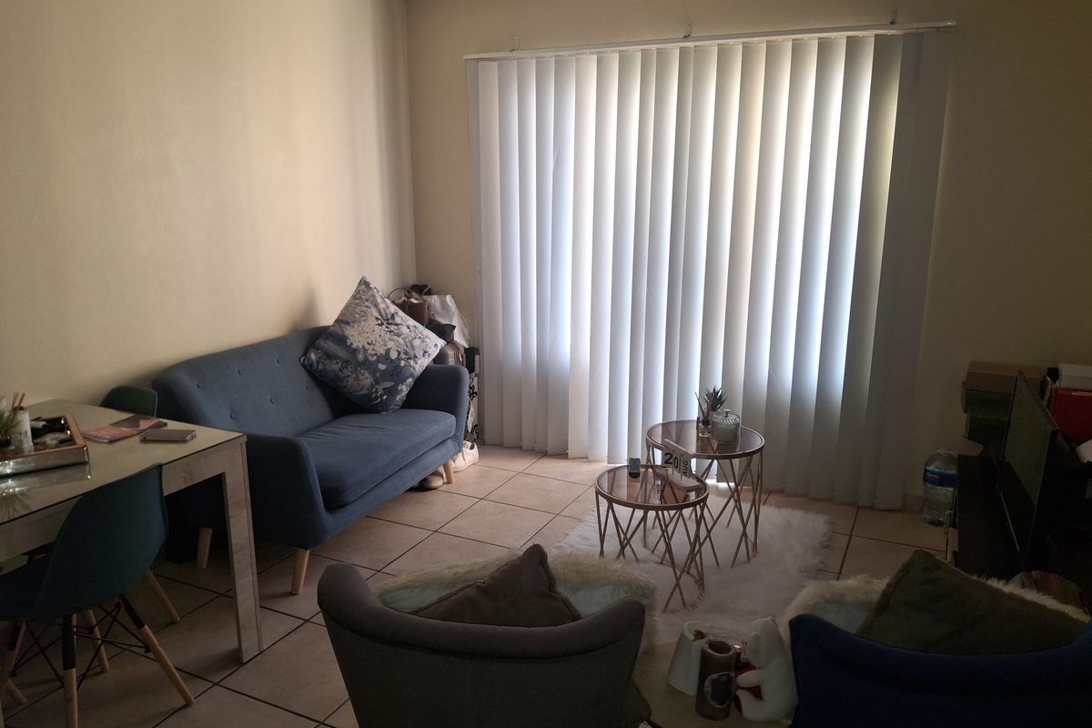 A living room area featuring tiled flooring and vertical blinds covering a large window, providing a functional and neutral space.