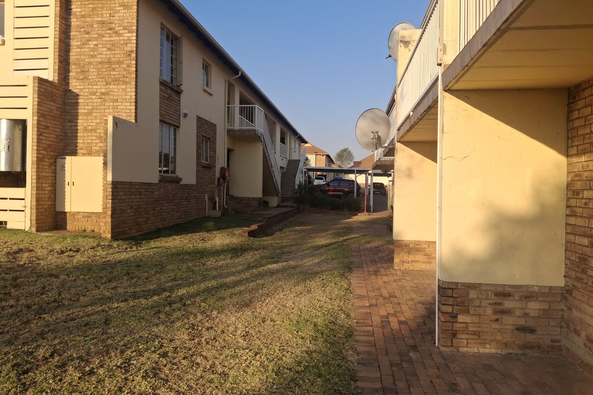 Exterior view of the apartment complex showing brick and plaster facade, paved walkways, and communal lawn area with balcony access.