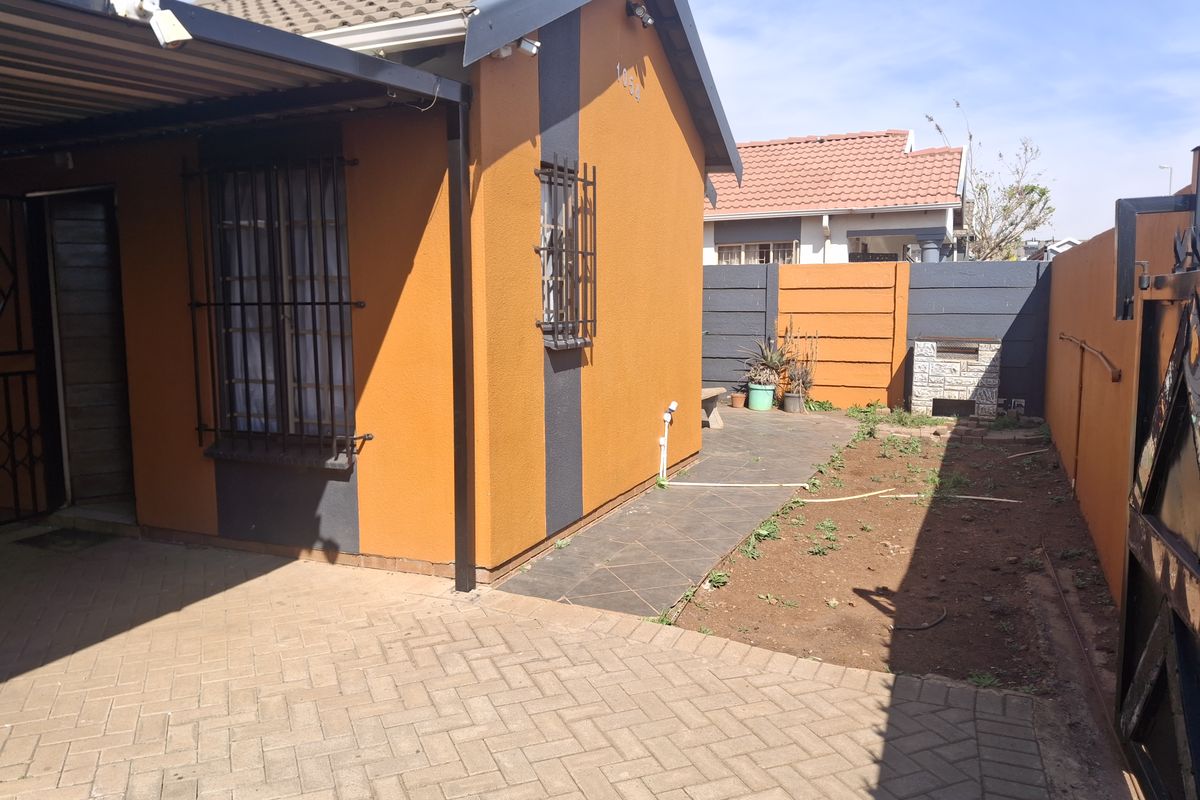 Rear exterior view of the property featuring a paved patio area, built-in braai, and a small garden space enclosed by boundary walls.