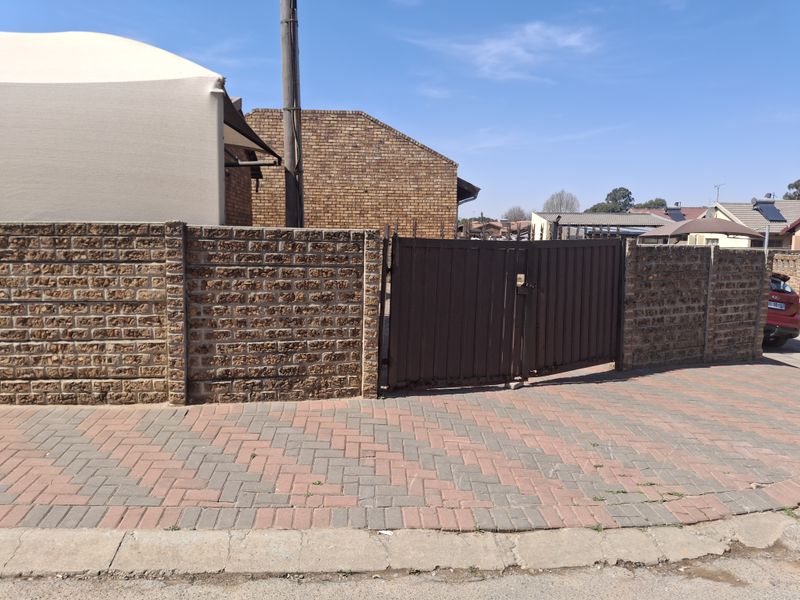 Front view of the property featuring a brick boundary wall, metal entrance gate, and paved driveway area.