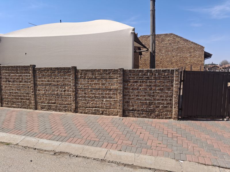 Street view showing a brick boundary wall, paved sidewalk, and metal gate entrance to the property.