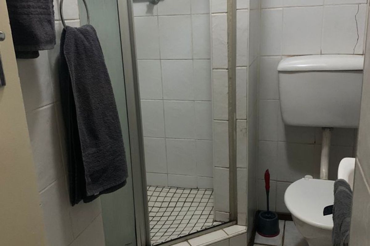 Compact bathroom featuring a shower enclosure and toilet with white tiled finishes, showing signs of wear and tile damage.
