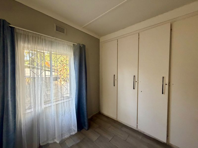 A functional bedroom featuring built-in cupboards, tiled flooring, and a window fitted with security bars.